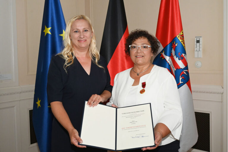 Zwei Frauen halten lächelnd eine Urkunde in der Hand, im Hintergrund Fahnen