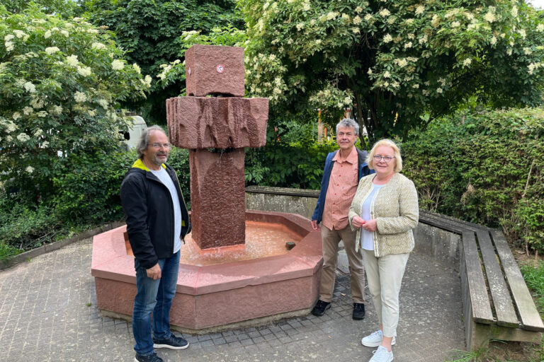drei Personen stehen vor einem Brunnen, Büsche und Bäume im Hintergrund