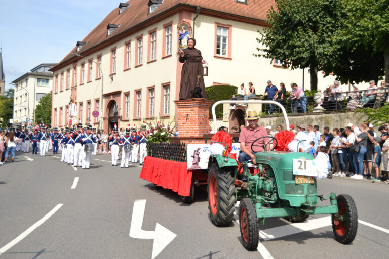 viele Personen am Straßenrand, Personen auf Straße verkleidet mit Instrumenten, Traktor mit einer verkleideten Frau auf einem Schornstein