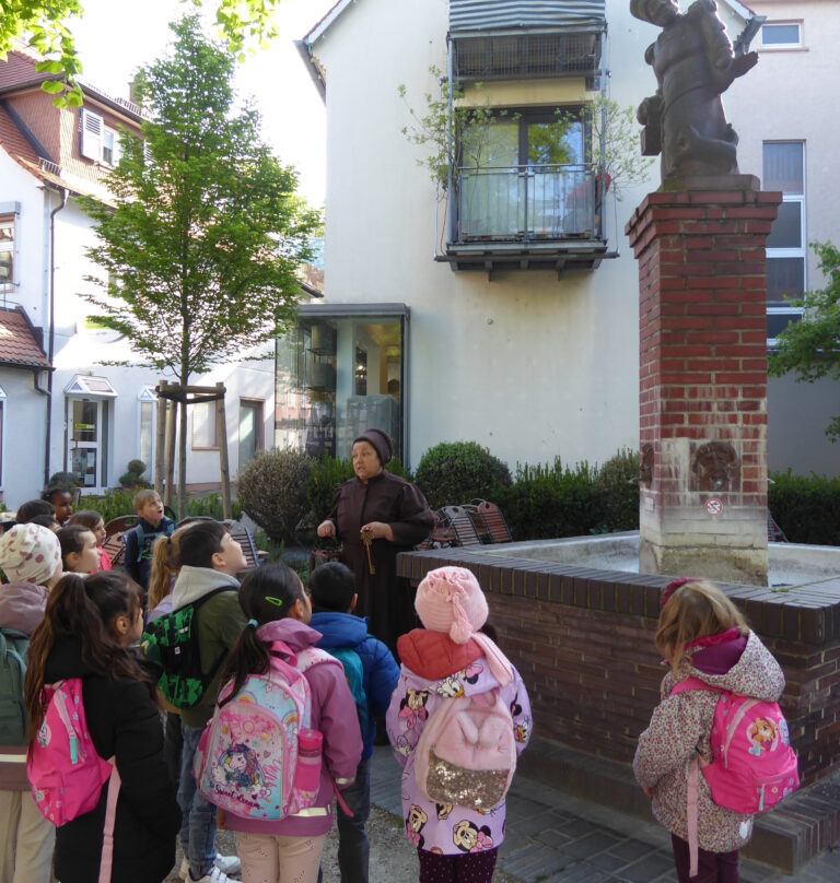 Kinder mit der Fraa vun Bensem vor Brunnen
