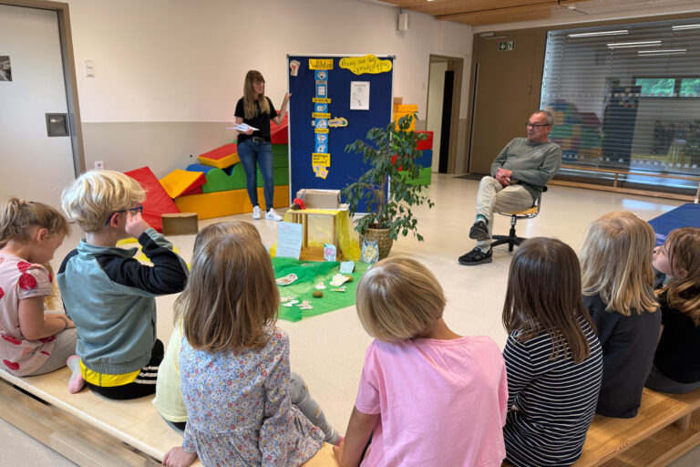 Kinder sitzen auf Bänken und schauen auf eine Stellwand, ein Mann sitzt im Hintergrund und eine Frau steht an der Stellwand