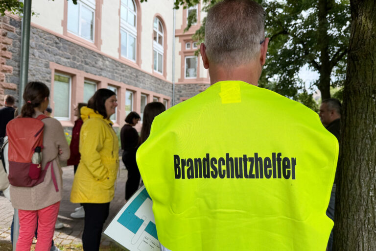 Mann mit gelber Weste mit der Aufschrift Brandschutzhelfer im Vordergrund und im Hintergrund weitere Personen